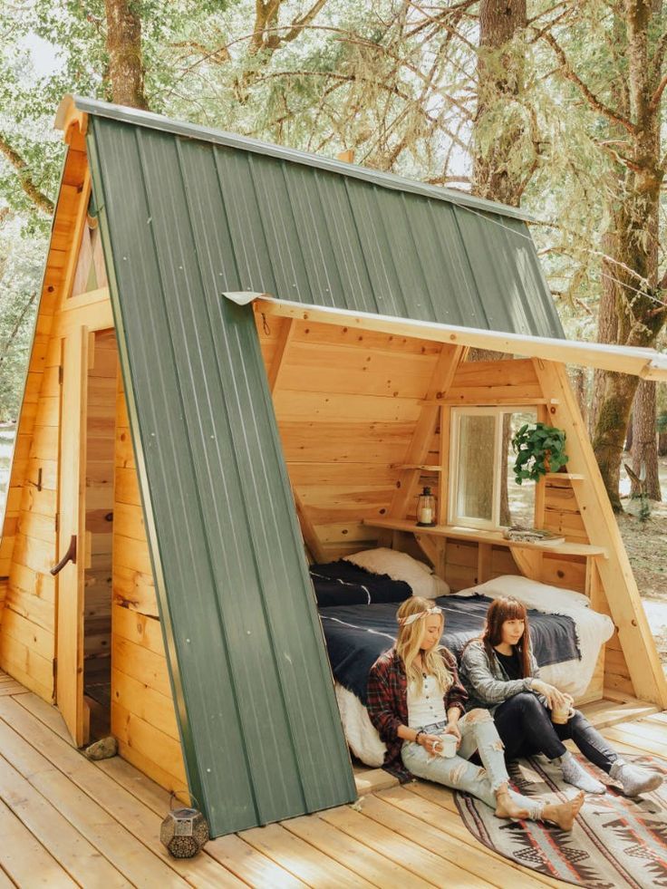 Duas pessoas sentadas na entrada de um mini chalé A-frame de madeira
